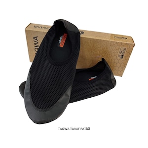 May include: Black mesh slip-on shoes with a rounded toe and a black leather-like trim. The shoes are displayed with a cardboard box in the background. The text "TAQWA" is visible on the insole and the box. The text "TAQWA TAVAF PATIĞI" is at the bottom.