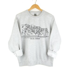 May include: A light gray sweatshirt with a black and white line drawing of a mountain range and the text "KAUAI, HAWAII". The sweatshirt is hanging on a wooden hanger against a white background.