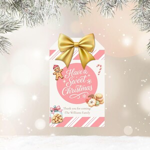 May include: A Christmas gift tag with a gold bow at the top. The tag has a pink and white striped background and features a gingerbread man, candy cane, and the text "Have a Sweet Christmas". A jar of cookies and a stack of cookies are also shown.