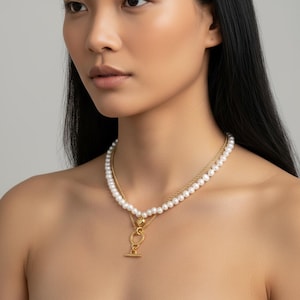 Puede incluir: Un collar de dos capas con perlas blancas y una cadena dorada. El collar tiene un colgante dorado con un cierre de palanca. La joya la lleva una persona.