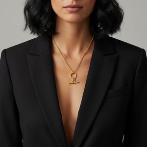 Op de afbeelding: Een goudkleurige ketting met een cirkelvormige hanger en een horizontale balk. De ketting wordt gedragen over een zwarte blazer. De hanger hangt aan een fijne ketting, wat een eenvoudige maar elegante uitstraling creëert. De sieraden zijn een klassiek accessoire.