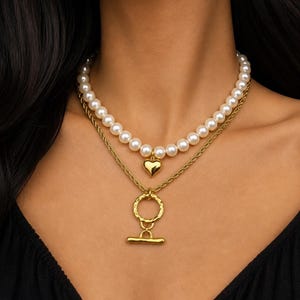 May include: A layered necklace featuring a strand of faux pearls and a gold chain. The top layer has a heart pendant, while the bottom layer has a textured circle and bar clasp. The jewelry is gold-toned.
