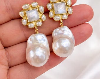 May include: A pair of elegant gold-toned earrings. Each earring features a square, iridescent center stone surrounded by small pearls. A large, baroque pearl dangles below. The earrings are held in a hand against a white background.