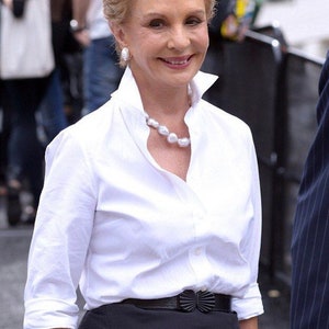 May include: A crisp white button-down shirt with a pointed collar and three-quarter length sleeves, paired with a black skirt featuring a black belt with a decorative bow-shaped buckle, accented by a large white pearl necklace and pearl earrings.