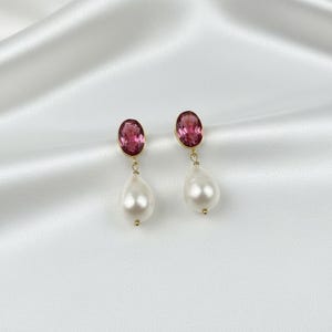 May include: A pair of gold earrings with oval, pink gemstones and teardrop-shaped white pearls. The earrings hang from a gold hook and are set against a white, silky background.