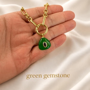 May include: A gold-toned chain necklace featuring a large circular link and a teardrop-shaped green pendant with a small dark oval stone in the center. The text "green gemstone" is printed in gold at the bottom of the image.
