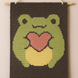 Può includere: Una rana verde all'uncinetto che tiene un cuore rosa, appesa a un muro con un'asta di legno e uno spago bianco.