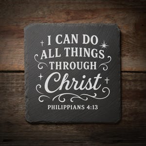 May include: A square, dark gray slate coaster with the white text "I CAN DO ALL THINGS THROUGH Christ" and "PHILIPPIANS 4:13". The coaster has decorative flourishes and is set against a wooden background.