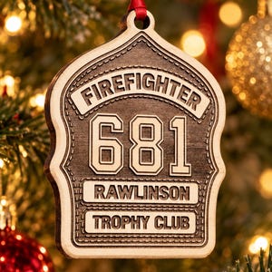May include: A wooden firefighter helmet-shaped ornament with a dark brown finish and light wood edges. It features the engraved text FIREFIGHTER, 681, RAWLINSON, and TROPHY CLUB. The ornament hangs from a red ribbon loop at the top center.