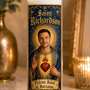 May include: A tall cylindrical glass prayer candle featuring a white wax pillar. The label displays a portrait of a man with a halo, wearing blue robes, with the text Saint Richardson and Patron Saint of Sarcasm printed in a decorative font.