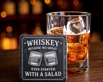 May include: A square, black slate coaster with white text that reads "WHISKEY BECAUSE NO GREAT EVER STARTED WITH A SALAD." A rocks glass with ice and amber liquid sits next to the coaster. The background is blurred with warm lighting.