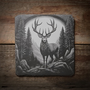 以下が含まれることがあります： 森の中に立つ大きな角を持つ雄鹿の、詳細な白黒イラストが描かれた正方形のスレートコースター。アートワークには、木、岩、アーチ型の背景が含まれています。