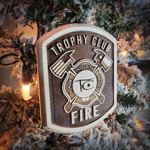 May include: Wooden plaque with the words "Trophy Club Fire" and a fire department emblem. The emblem includes an American flag, a Texas flag, axes, and the words "Honor, Integrity, Pride". The plaque is brown and tan.