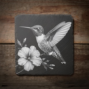 May include: A square, black slate coaster featuring a detailed, grayscale illustration of a hummingbird with outstretched wings, perched near a large hibiscus flower. The coaster is set against a rustic wooden background.