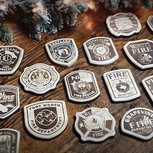 May include: A collection of wooden ornaments featuring various fire department emblems. Each ornament is a light brown color with dark brown engraved details, including text and symbols. The ornaments are arranged on a wooden surface, with a festive evergreen branch in the background.