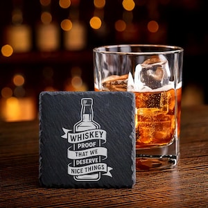 May include: A square, black slate coaster with white text and a whiskey bottle graphic. The text reads "Whiskey Proof That We Deserve Nice Things." A glass of whiskey with ice sits next to the coaster on a wooden surface.