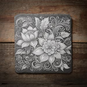 May include: Square, dark grey slate coaster with a detailed floral design. The design features large, stylized flowers, leaves, and swirling patterns in white. The coaster sits on a wooden surface, creating a high-contrast decorative piece.