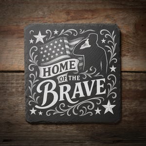 May include: A square, dark gray slate coaster with white etched design. The design includes the American flag, a saluting silhouette, stars, and the words "HOME OF THE BRAVE" in a decorative font. The coaster is on a wooden surface.