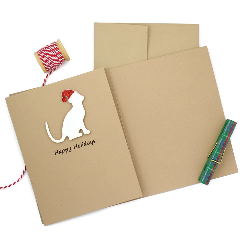 May include: A set of holiday greeting cards with a white cat silhouette wearing a Santa hat. The cards are tan with the words "Happy Holidays" printed below the cat. A spool of red and white twine and a plaid clip are also in the image.