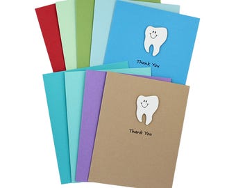 May include: A collection of rectangular greeting cards in various colors including red, green, blue, purple, and tan. Each card features a white die-cut tooth shape with a smiling face and the printed text Thank You in black ink.