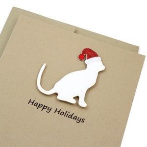 May include: A holiday greeting card featuring a white cat silhouette wearing a red Santa hat with a white trim and a red pom-pom. The words "Happy Holidays" are printed in black on a tan card.
