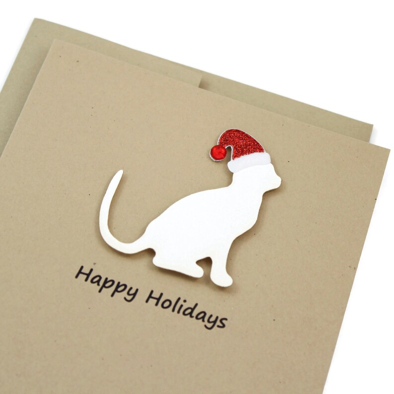 May include: A holiday greeting card featuring a white cat silhouette wearing a red Santa hat with a white trim and a red pom-pom. The words "Happy Holidays" are printed in black on a tan card.