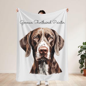 May include: A white blanket with a detailed illustration of a German Shorthaired Pointer dog. The dog has brown and white fur, with a focused gaze. The words "German Shorthaired Pointer" are written in a script font across the top.
