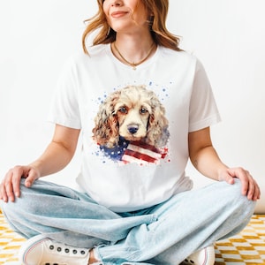 May include: White t-shirt featuring a watercolor illustration of a Cocker Spaniel with an American flag. The dog has brown and cream fur, with red, white, and blue accents. The shirt is a classic crew neck style.