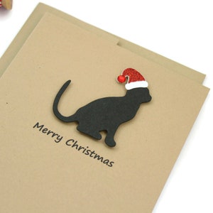 May include: A Christmas card featuring a black cat silhouette wearing a red Santa hat with a white trim and a red pom-pom. The words "Merry Christmas" are printed in black below the cat on a tan card.