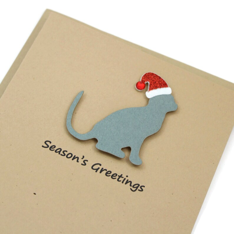 May include: A Christmas card featuring a gray cat silhouette wearing a red glitter Santa hat. The words "Season's Greetings" are printed in black below the cat on a tan card.
