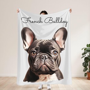 May include: A white blanket featuring a detailed illustration of a French Bulldog's face. The dog is black with brown eyes and white markings. The words "French Bulldog" are written in a cursive font across the top.