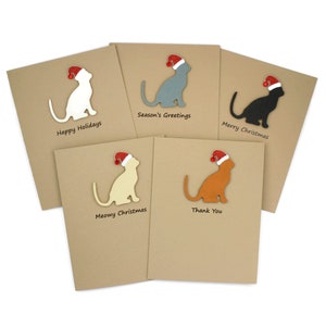 May include: Five greeting cards with cat silhouettes wearing Santa hats. The cards are various colors: white, gray, black, cream, and orange. Each card has a different holiday message: Happy Holidays, Season's Greetings, Merry Christmas, Meowy Christmas, and Thank You.
