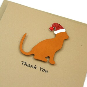 May include: A brown cat silhouette wearing a red and white Santa hat is affixed to a tan card. The words "Thank You" are printed in black below the cat. The card is a festive design.