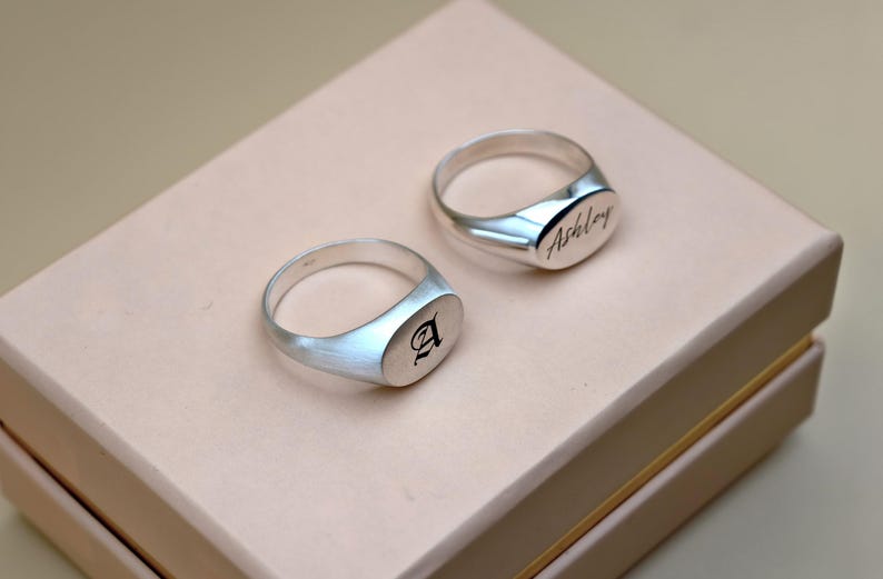 Puede incluir: Dos anillos de sello de plata en una caja rosa claro. Un anillo tiene la letra "A" grabada y el otro anillo tiene el nombre "Ashley" grabado.