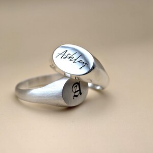 Puede incluir: Dos anillos de sello de plata. Un anillo tiene el nombre "Ashley" grabado en la parte superior. El otro anillo tiene la letra "A" grabada en la parte superior.