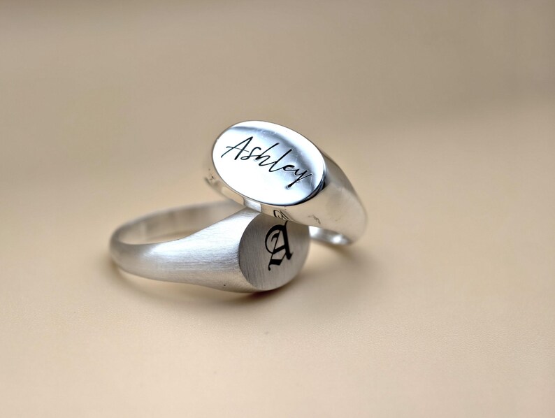 Puede incluir: Dos anillos de sello de plata. Un anillo tiene el nombre "Ashley" grabado en la parte superior. El otro anillo tiene la letra "A" grabada en la parte superior.