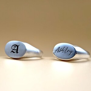 Puede incluir: Dos anillos de sello de plata con tapas ovaladas. El anillo de la izquierda tiene la letra "A" grabada en una fuente g&oacute;tica. El anillo de la derecha tiene el nombre "Ashley" grabado en cursiva.