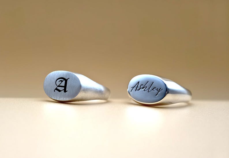 Puede incluir: Dos anillos de sello de plata con tapas ovaladas. El anillo de la izquierda tiene la letra "A" grabada en una fuente g&oacute;tica. El anillo de la derecha tiene el nombre "Ashley" grabado en cursiva.