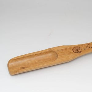 May include: A wooden scoop with a handle, made of bamboo. The scoop has a small, rounded depression in the front. The handle is engraved with the words "Living Roots" and a tree design.
