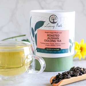 May include: A clear glass teacup with a handle, filled with golden tea, sits next to a cylindrical container of Living Roots Roasted Alishan Oolong Tea. The container has a white top and a green base, with text and a logo. Loose tea leaves are on a wooden scoop.