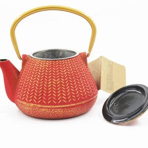 May include: Red cast iron teapot with a gold handle and a matching lid. The teapot has a textured pattern and a built-in infuser. A small tag is attached to the teapot.