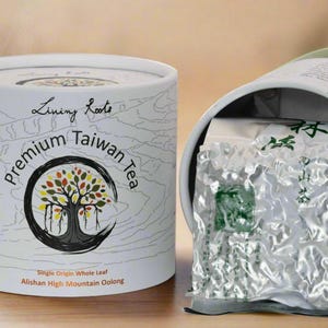 May include: A white cylindrical container of "Premium Taiwan Tea" with a tree logo. The container is open, revealing a foil package of tea. Text on the container includes "Living Roots", "Single Origin Whole Leaf", and "Alishan High Mountain Oolong".