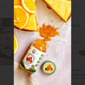 May include: An overhead shot features a jar and a small tin of Lakadong Turmeric, with a slice of cake and orange slices. The jar is open, spilling turmeric powder. The labels display the product name and a graphic of turmeric roots.
