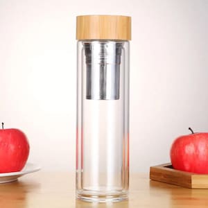 May include: A clear glass water bottle with a bamboo lid and a stainless steel tea infuser. The bottle is sitting on a wooden surface.