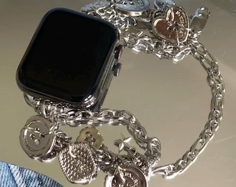 May include: A black smartwatch with a silver chain band. The band features various charms, including buttons, hearts, and stars. The watch and band are displayed on a reflective surface, with a denim background.