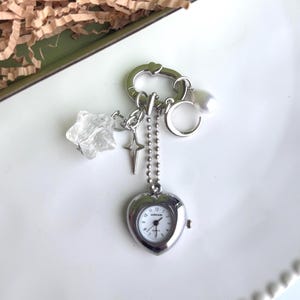 May include: A silver-toned heart-shaped watch with a white face and black numerals, attached to a keychain with charms including a star, a cross, a crescent moon, and a clear crystal star. The watch is on a white plate.