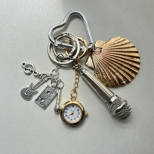 Może przedstawiać: Srebrny brelok do kluczy z sercowatym kółkiem, zawieszką w kształcie muszli, zawieszką w kształcie mikrofonu, zawieszką w kształcie gitary, zawieszką w kształcie kasety i małą zawieszką w kształcie zegarka.