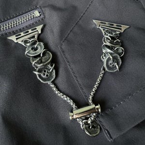 May include: Silver-toned Apple Watch band featuring a unique design. The band incorporates decorative, textured buttons and chain links. The band is shown against a dark gray fabric background, with a zipper visible in the upper left corner.