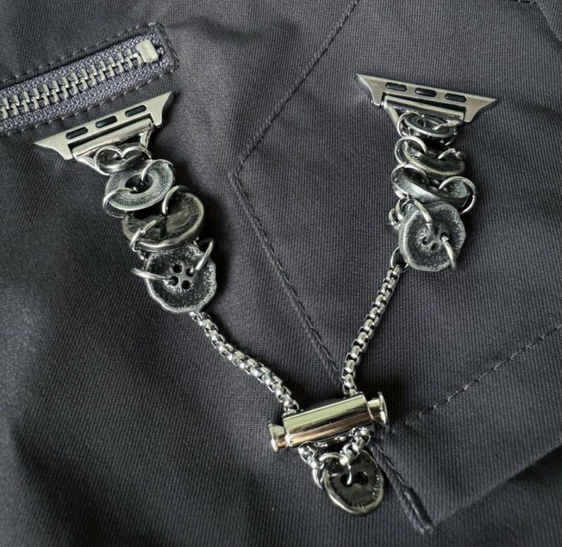 May include: Silver-toned Apple Watch band featuring a unique design. The band incorporates decorative, textured buttons and chain links. The band is shown against a dark gray fabric background, with a zipper visible in the upper left corner.