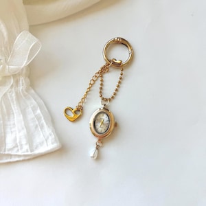 May include: A gold-toned oval watch pendant attached to a circular keyring by a beaded chain. The watch face is white with gold hands and markers. A small heart-shaped charm hangs from a separate gold chain, and a white teardrop pearl hangs below.
